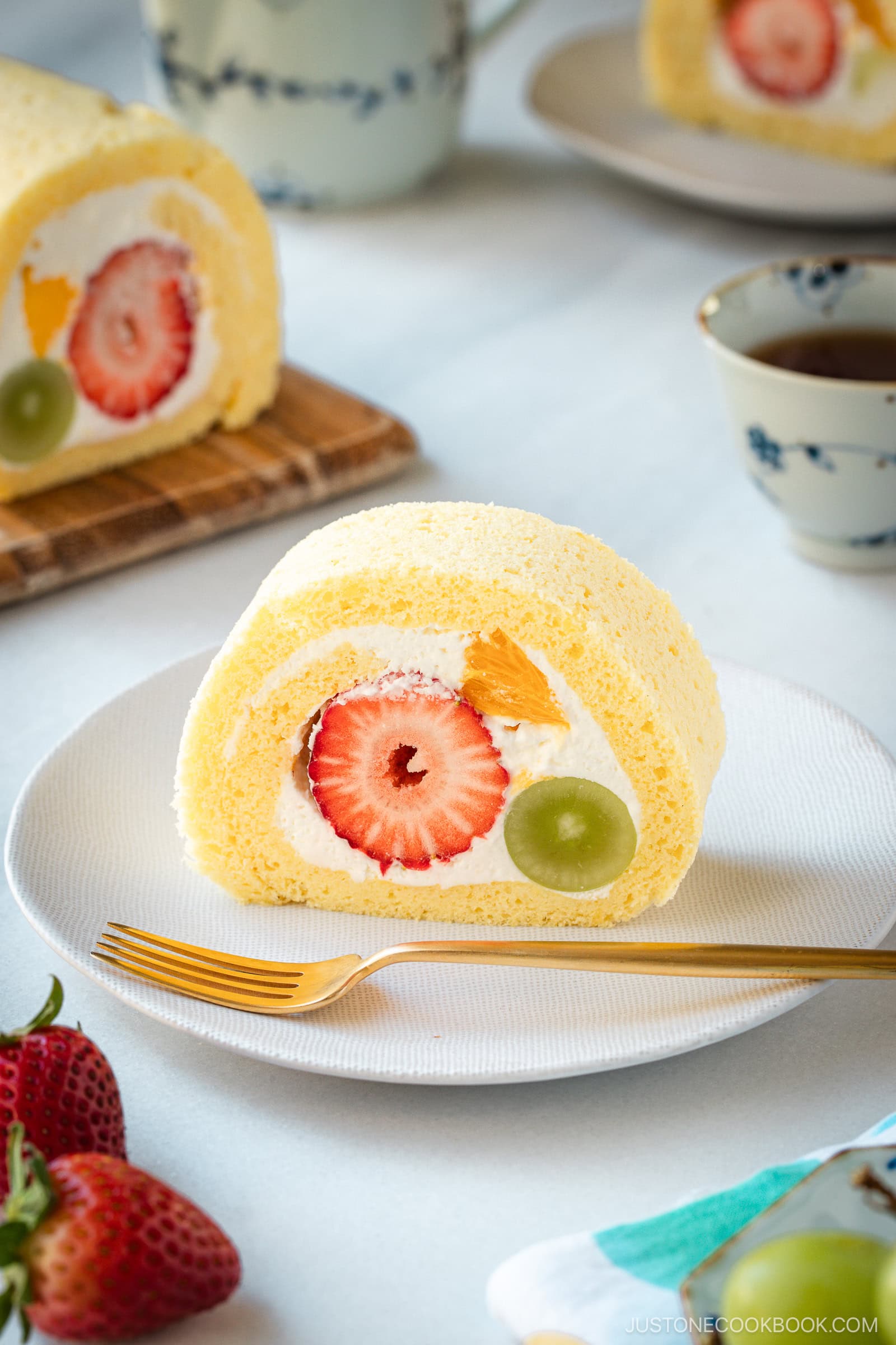 A slice of Japanese fruit roll cake filled with whipped cream, strawberries, orange pieces, and a grape, served on a white plate with a gold fork. Whole strawberries and a teacup are nearby.