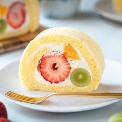 A slice of fluffy yellow Swiss roll cake filled with whipped cream, a strawberry, a green grape, and a piece of orange sits on a white plate with a gold fork. Whole strawberries and another cake slice are in the background.