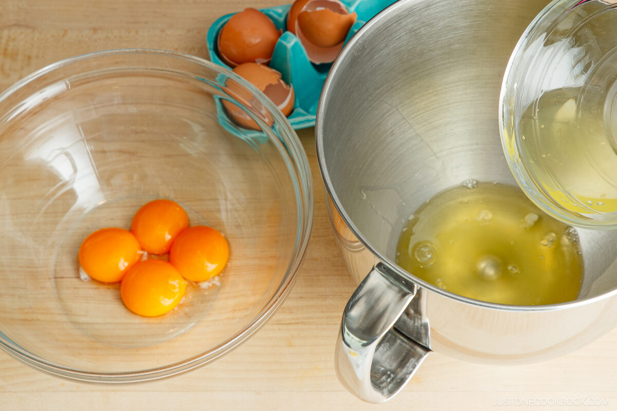 A glass bowl with four egg yolks, a metal mixing bowl with egg whites being poured in, and a container holding cracked eggshells sit on a wooden surface.