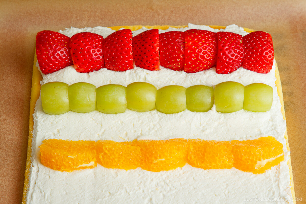 A rectangular cake topped with three neat rows of sliced fruit: strawberries, green grapes, and orange segments, all placed on a layer of white frosting.
