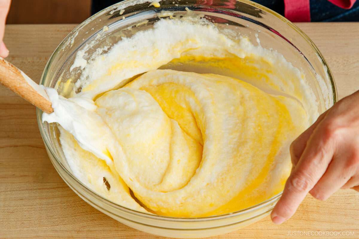 A person folds whipped egg whites into a yellow batter in a glass bowl using a spatula, preparing a mixture on a wooden surface.