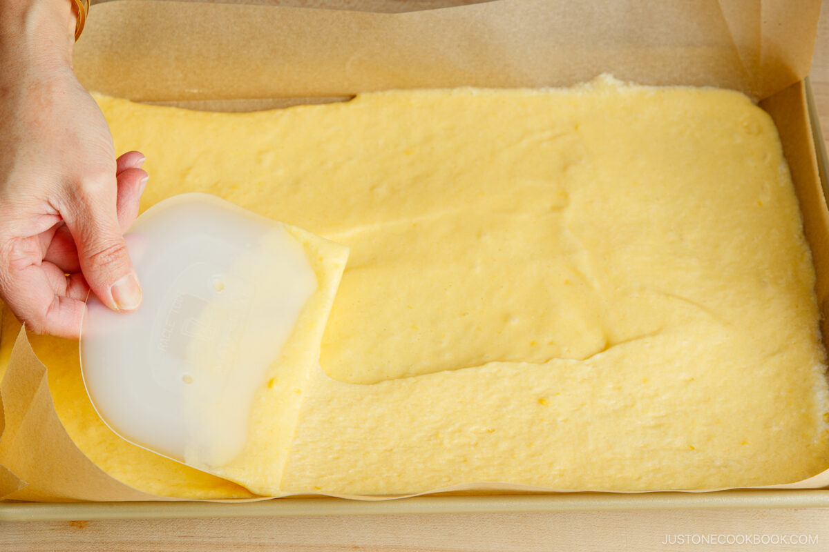 A hand uses a plastic scraper to spread yellow cake batter evenly in a parchment-lined baking pan.