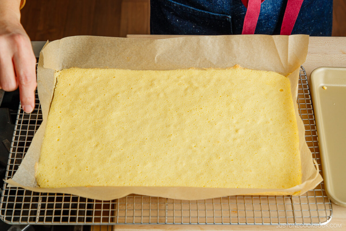 A person’s hand holds a baking tray lined with parchment paper and a thin, rectangular sponge cake, cooling on a wire rack atop a wooden surface.