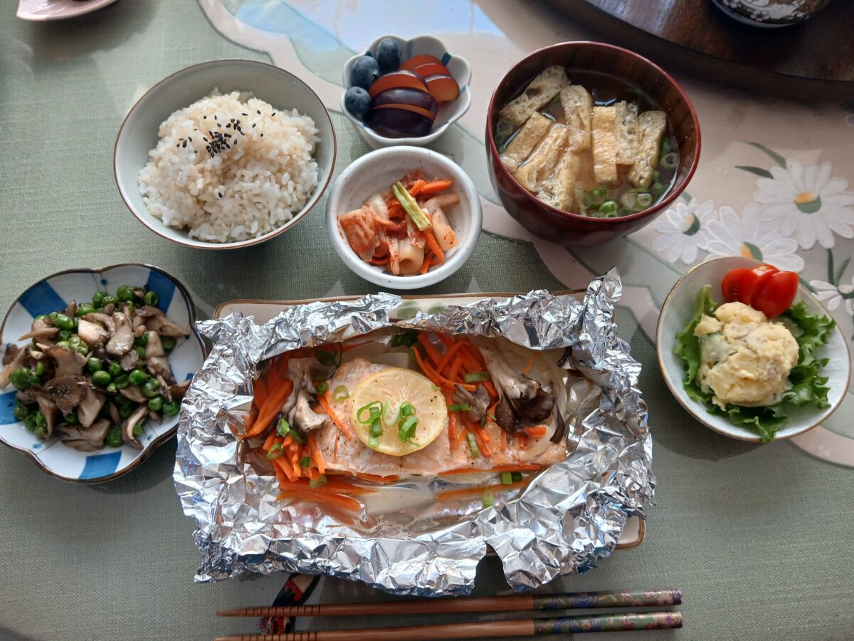 A Japanese meal with rice, miso soup, grilled salmon with lemon and vegetables in foil, sautéed mushrooms, potato salad, kimchi, and a small bowl of fruit, arranged neatly on a table with chopsticks.