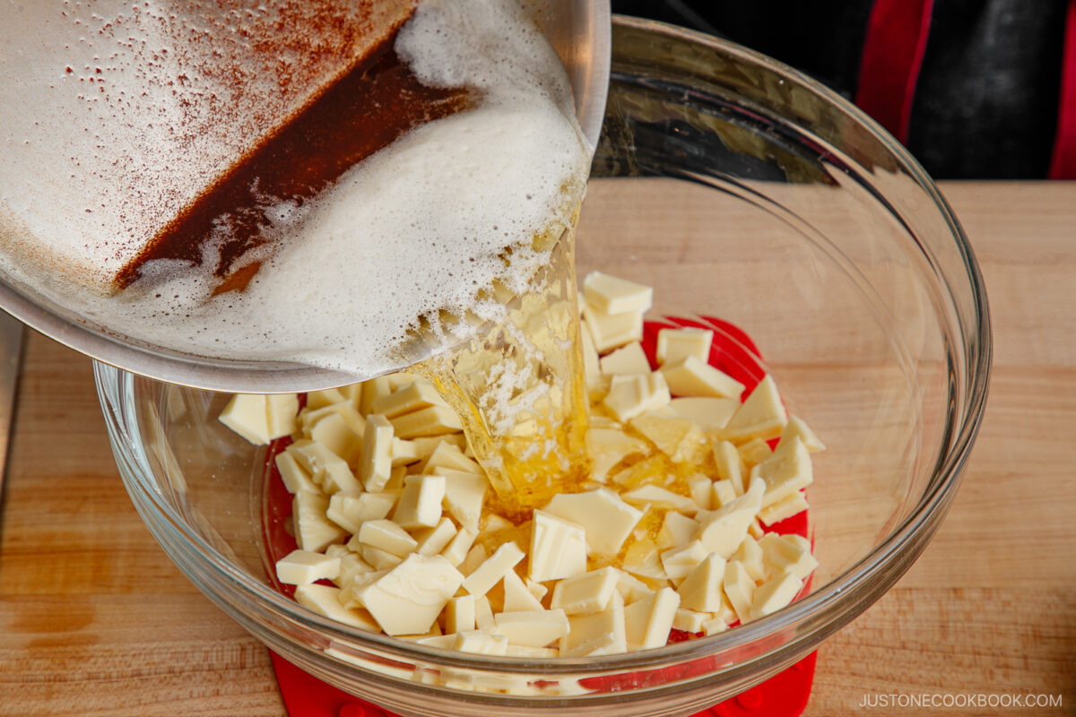 Browned butter is being poured from a metal saucepan into a glass bowl filled with chopped white chocolate pieces, likely to melt them together for a recipe. The bowl sits on a red silicone mat on a wooden surface.