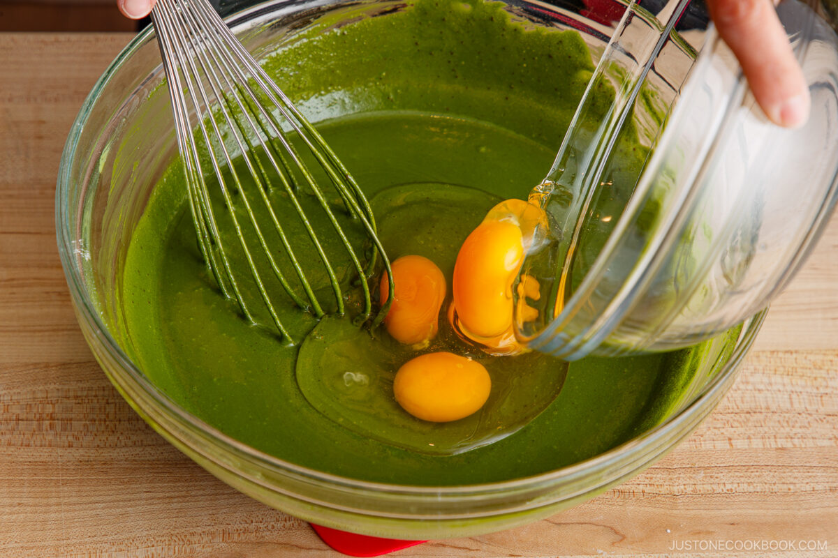 A hand pours three raw eggs from a glass bowl into a large bowl of green batter, while a metal whisk rests in the mixture on a wooden surface.