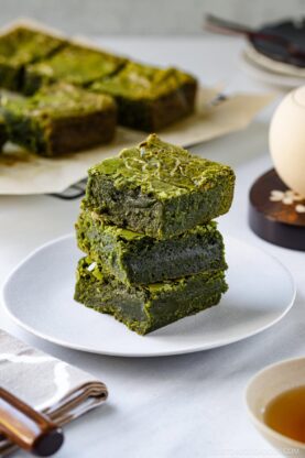 A stack of three green matcha brownies sits on a white plate, with more brownies in the background on parchment paper. A tea set and napkin are nearby on a light-colored table.