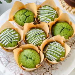 Seven matcha muffins in brown paper wrappers are arranged on a white plate. Four muffins have white icing drizzled on top, while the other three are plain. The scene is bright and inviting, with a hint of a coffee cup and utensils nearby.