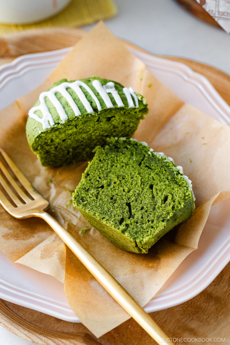 A sliced matcha muffin with white icing drizzle sits on brown parchment paper atop a pink plate. A gold fork rests beside the muffin.