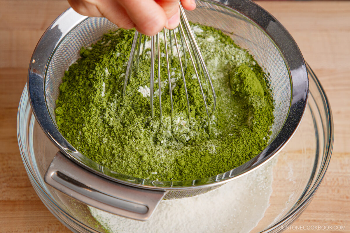 A hand uses a whisk to sift green matcha powder, flour, and other dry ingredients through a mesh sieve into a glass bowl on a wooden surface.