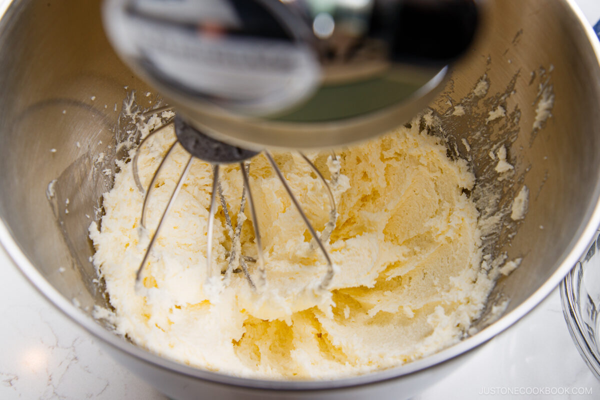 A stand mixer blends creamed butter and sugar in a metal mixing bowl, creating a light and fluffy mixture. The whisk attachment is partially covered with the mixture.