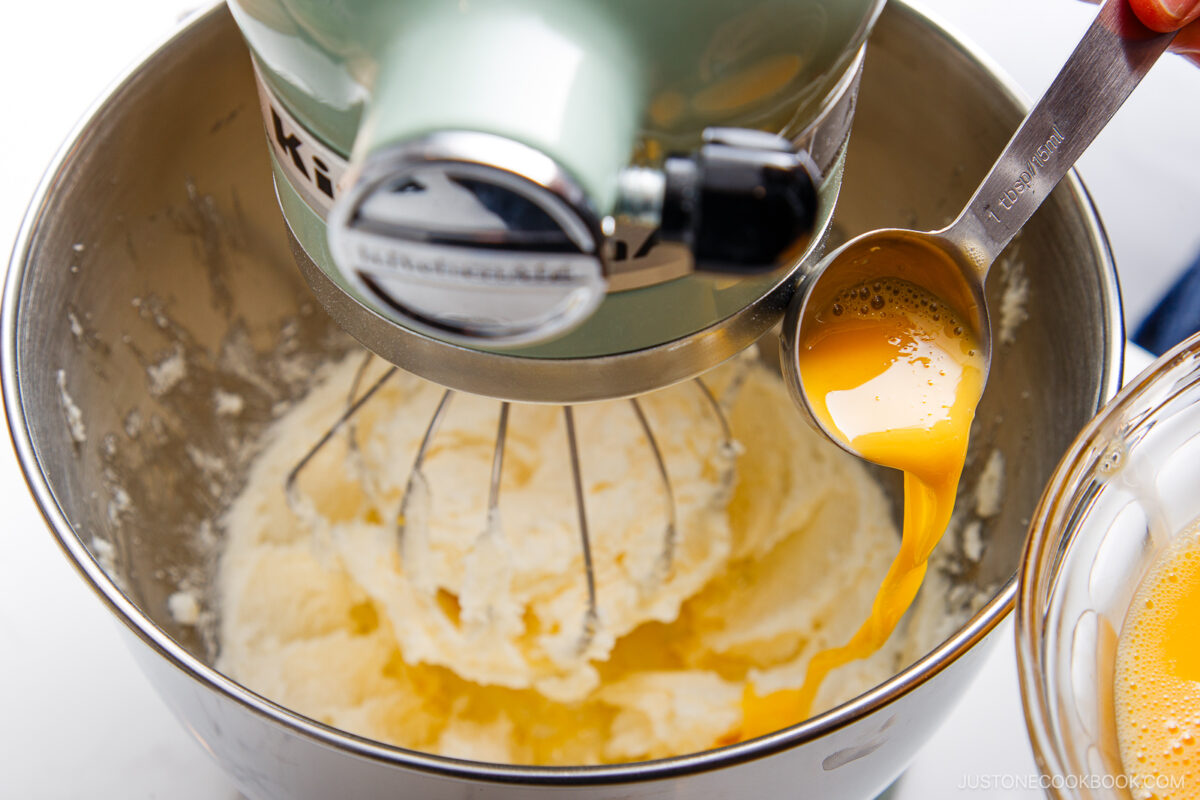 A close-up of a stand mixer beating a creamy mixture in its metal bowl while a person pours beaten eggs from a measuring spoon into the bowl.