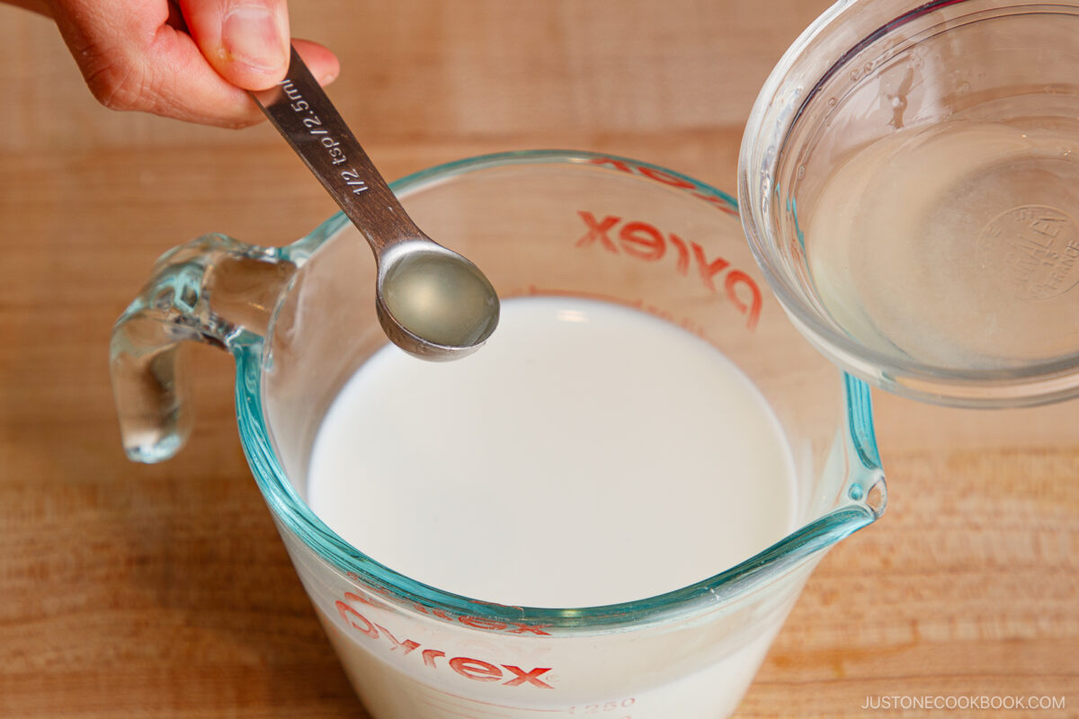 A hand holds a measuring spoon with liquid above a glass measuring cup filled with milk, preparing to add the liquid. Another hand holds a small bowl with more liquid next to the cup.