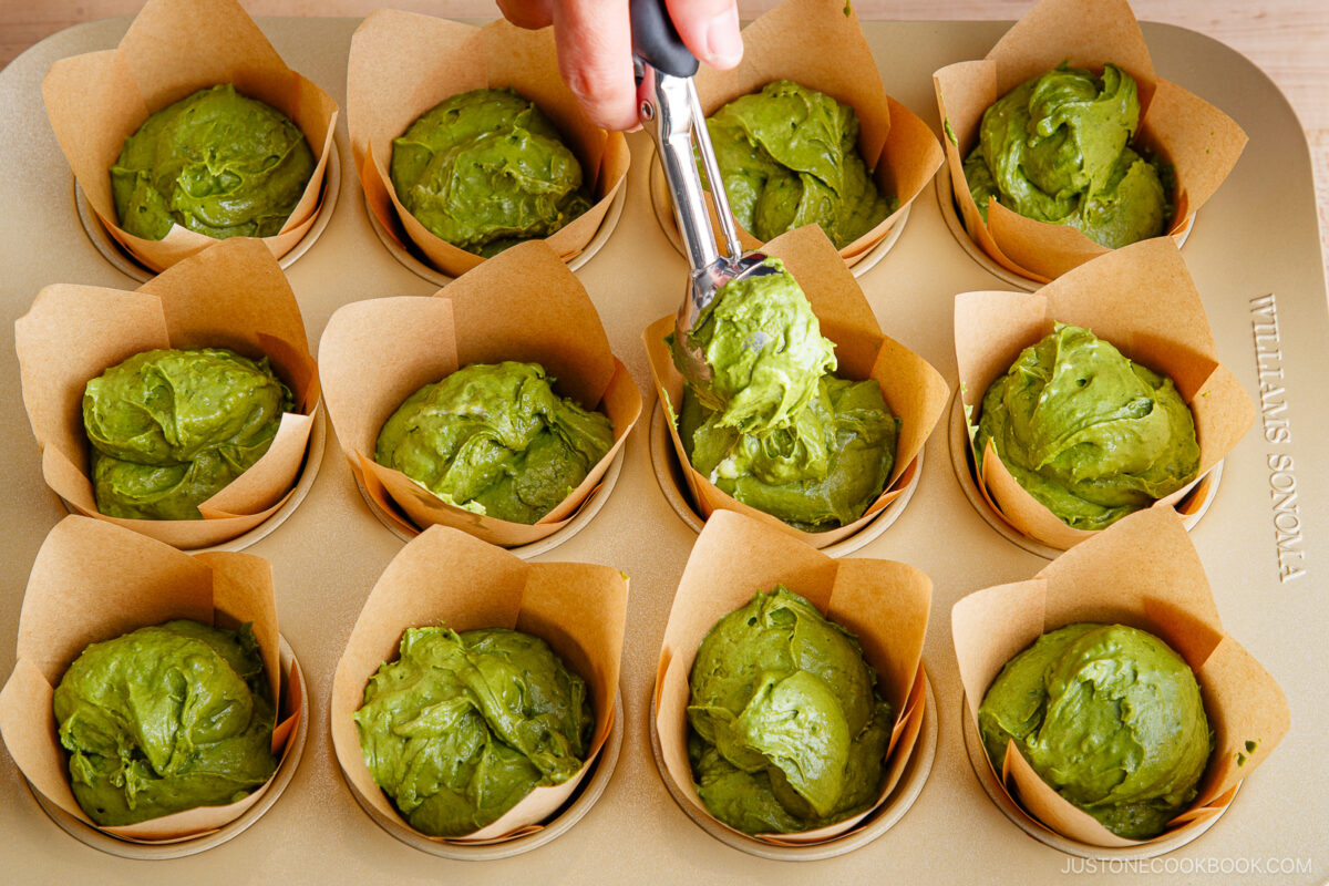 A hand uses an ice cream scoop to fill parchment-lined muffin cups in a baking tray with green matcha muffin batter.
