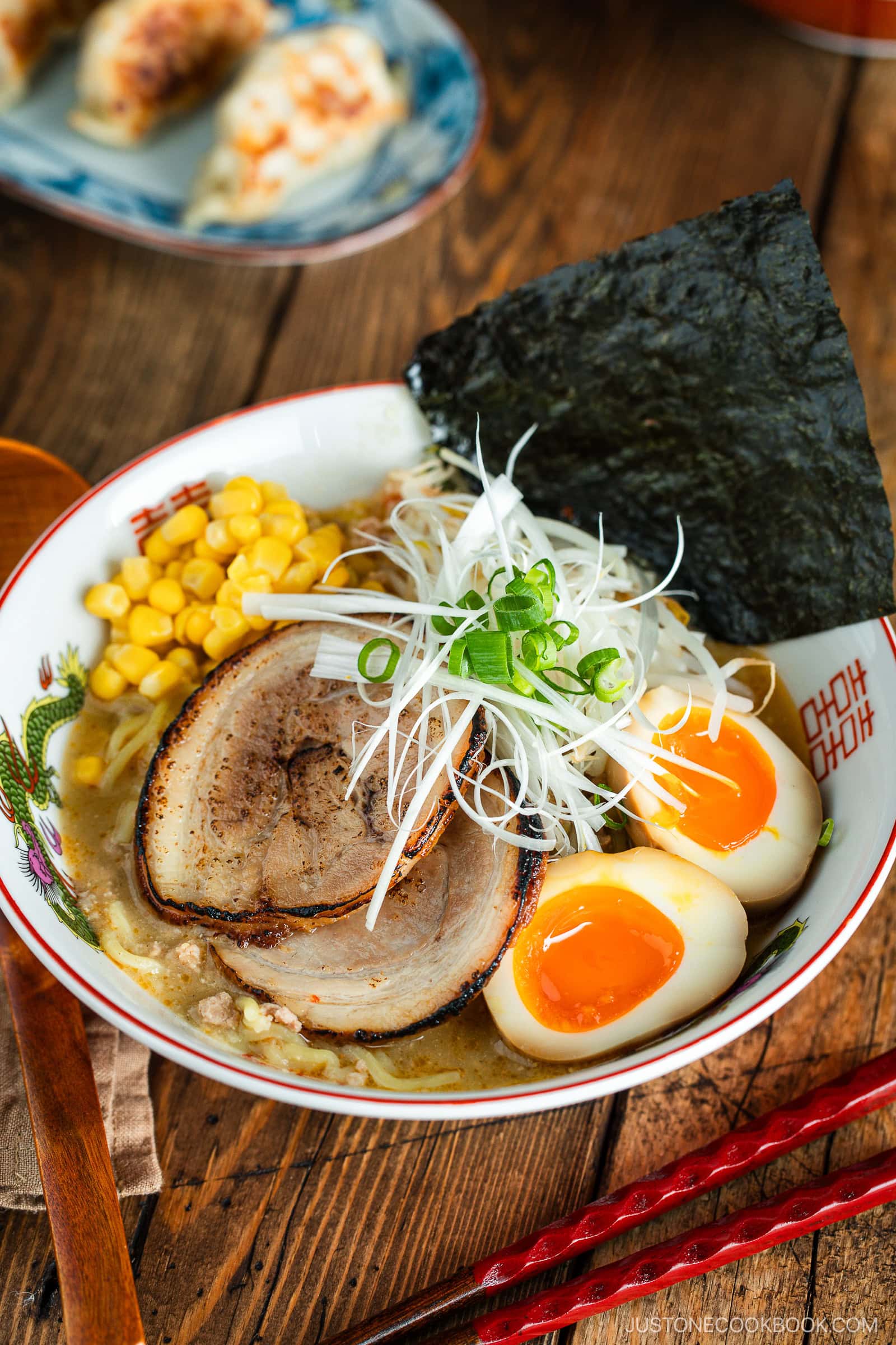 A bowl of ramen with sliced pork, soft-boiled eggs, corn, nori seaweed, green onions, and shredded white onions, served in a decorative bowl on a wooden table with red chopsticks.