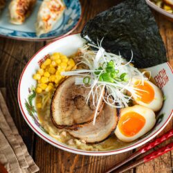 A bowl of ramen with sliced pork, soft-boiled eggs, corn, green onions, seaweed, and noodles in broth. Red chopsticks and a wooden spoon are nearby. Gyoza dumplings are in the background on a wooden table.