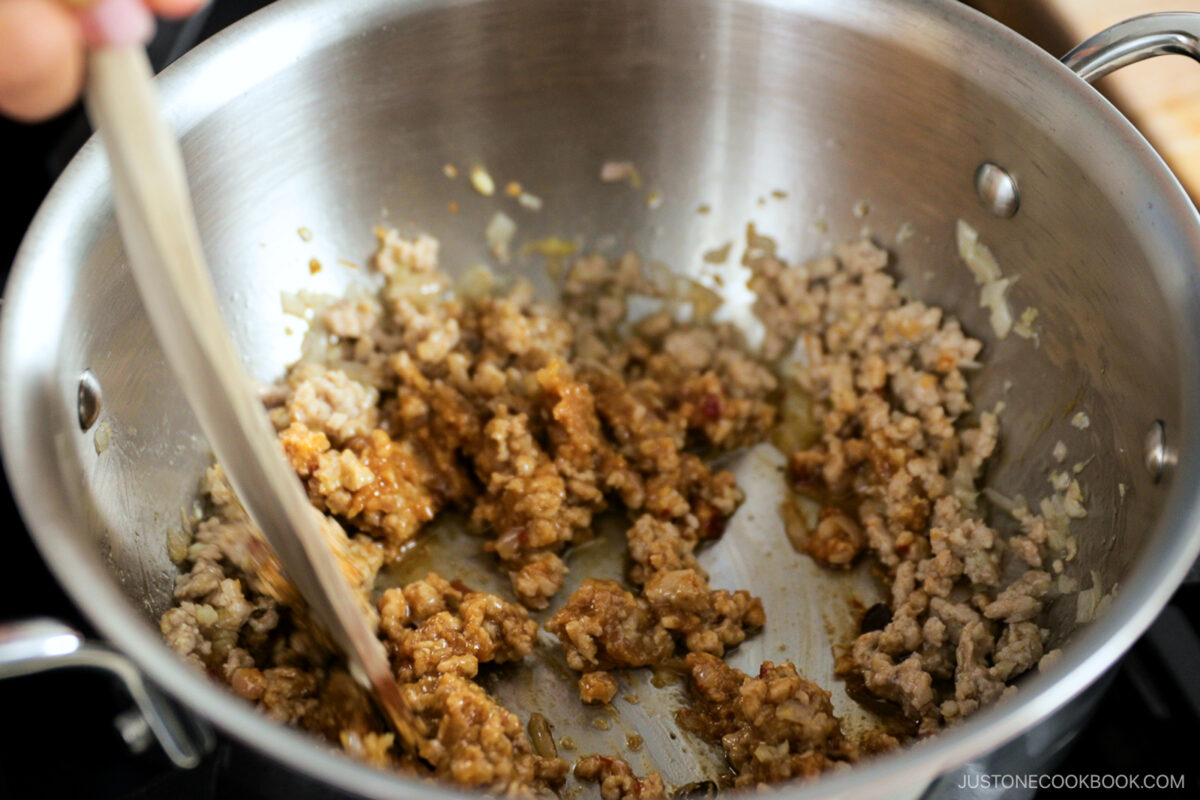 Ground meat is being cooked and stirred with a wooden spatula in a stainless steel pot on a stovetop. Some browned bits and savory sauce are visible in the mixture.