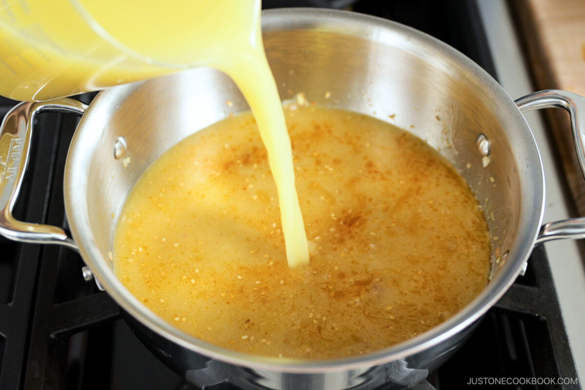 A close-up of yellow broth being poured from a measuring cup into a stainless steel pot with seasonings, on a stove.