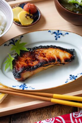 A piece of grilled fish with caramelized edges is served on a decorative white plate with a blue pattern, garnished with green leaves. Chopsticks and side dishes are partially visible around it.