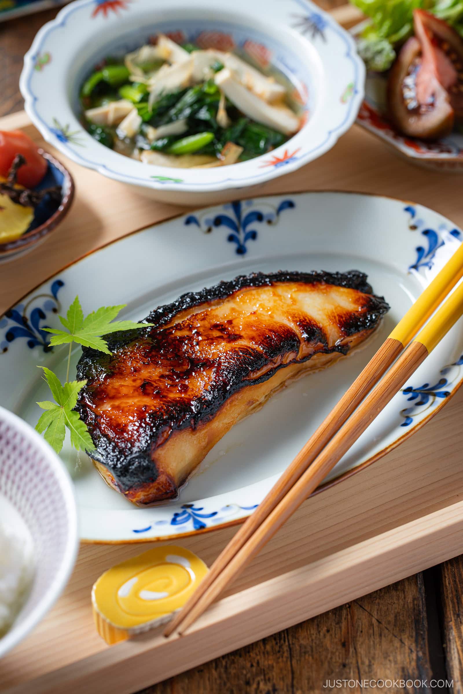 A beautifully plated serving of miso-glazed grilled fish garnished with green leaves, surrounded by small dishes of vegetables and rice, with chopsticks resting on the plate.