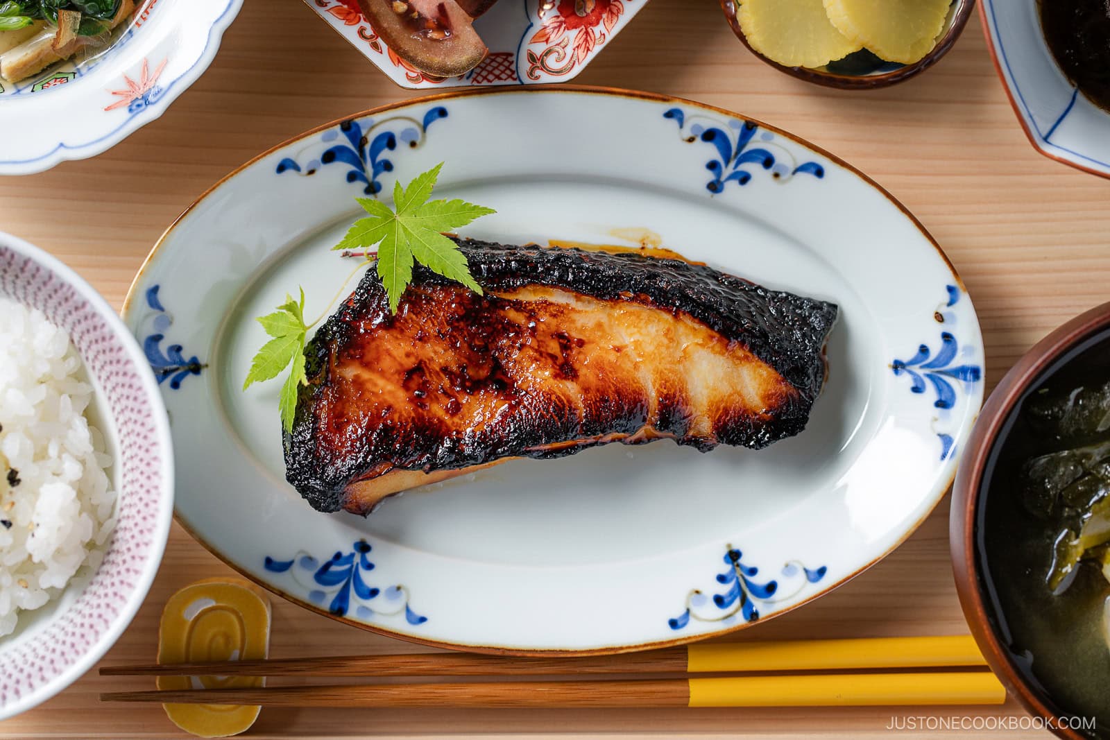 A grilled fillet of miso-glazed fish garnished with a green leaf, served on an oval plate with a blue floral pattern, surrounded by bowls of rice, pickles, and soup on a wooden table.