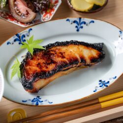 A plate with a glazed, grilled fish fillet garnished with a green leaf, surrounded by bowls of rice, pickles, soup, and salad, with chopsticks resting nearby on a wooden table.