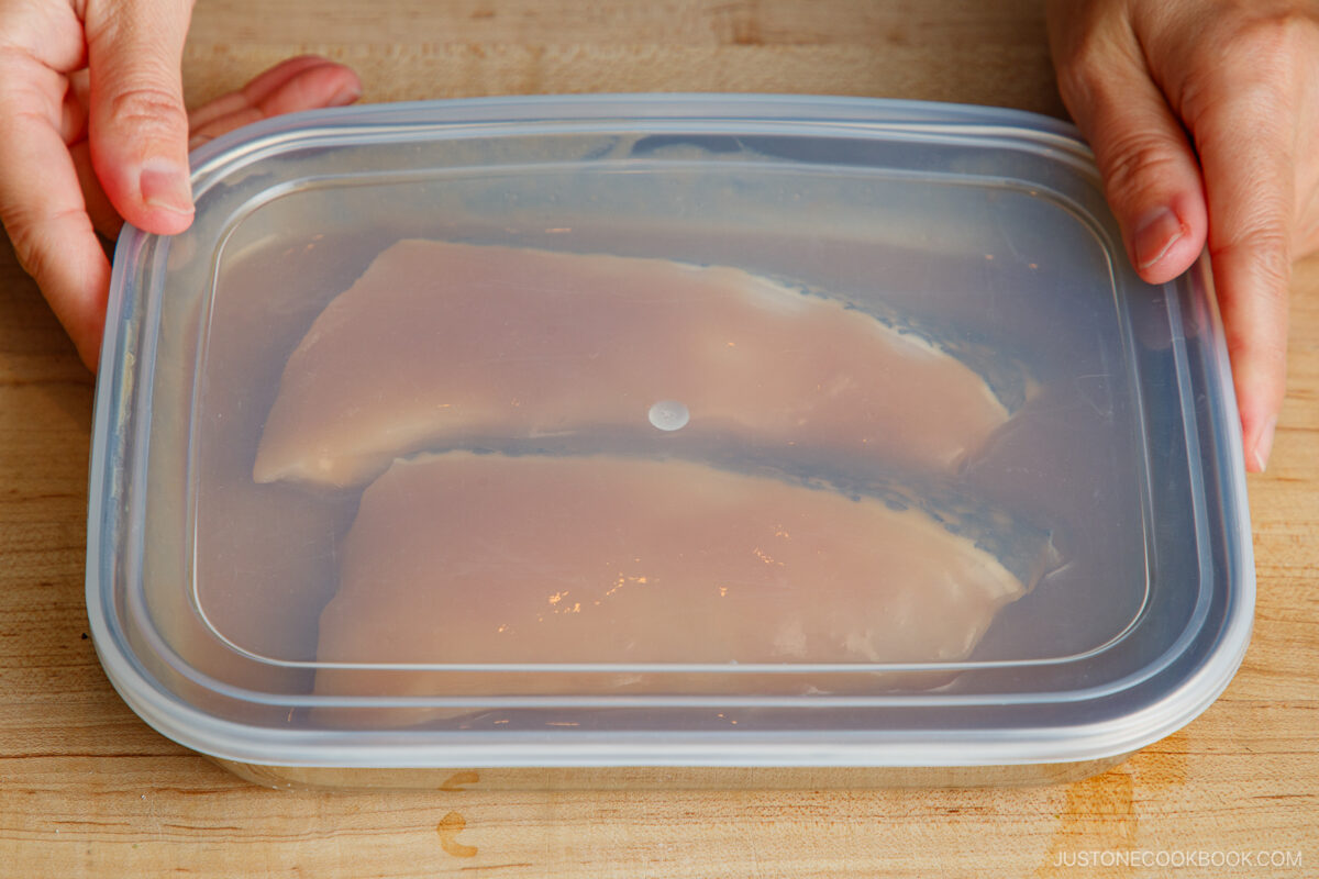 Two pieces of raw fish are marinating in a clear plastic container with a lid, placed on a wooden surface. Hands are holding the sides of the container.