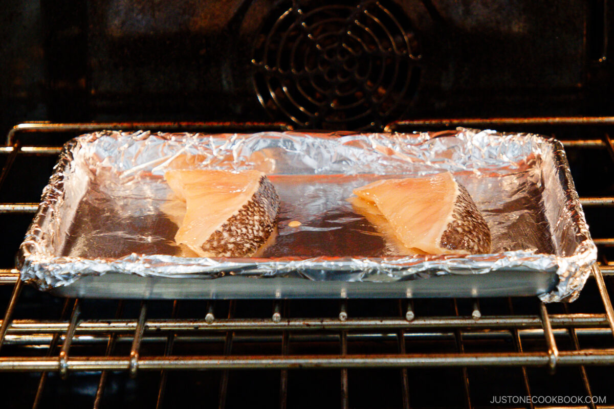 Two pieces of fish with skin on, placed on a foil-lined baking tray, are being cooked in an oven. The tray sits on the oven rack, and the oven interior is visible.
