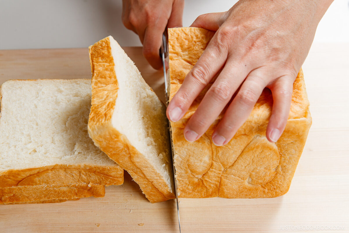 A person’s hands are slicing a loaf of fluffy, white bread on a wooden surface. Several slices are already cut and stacked to the side.
