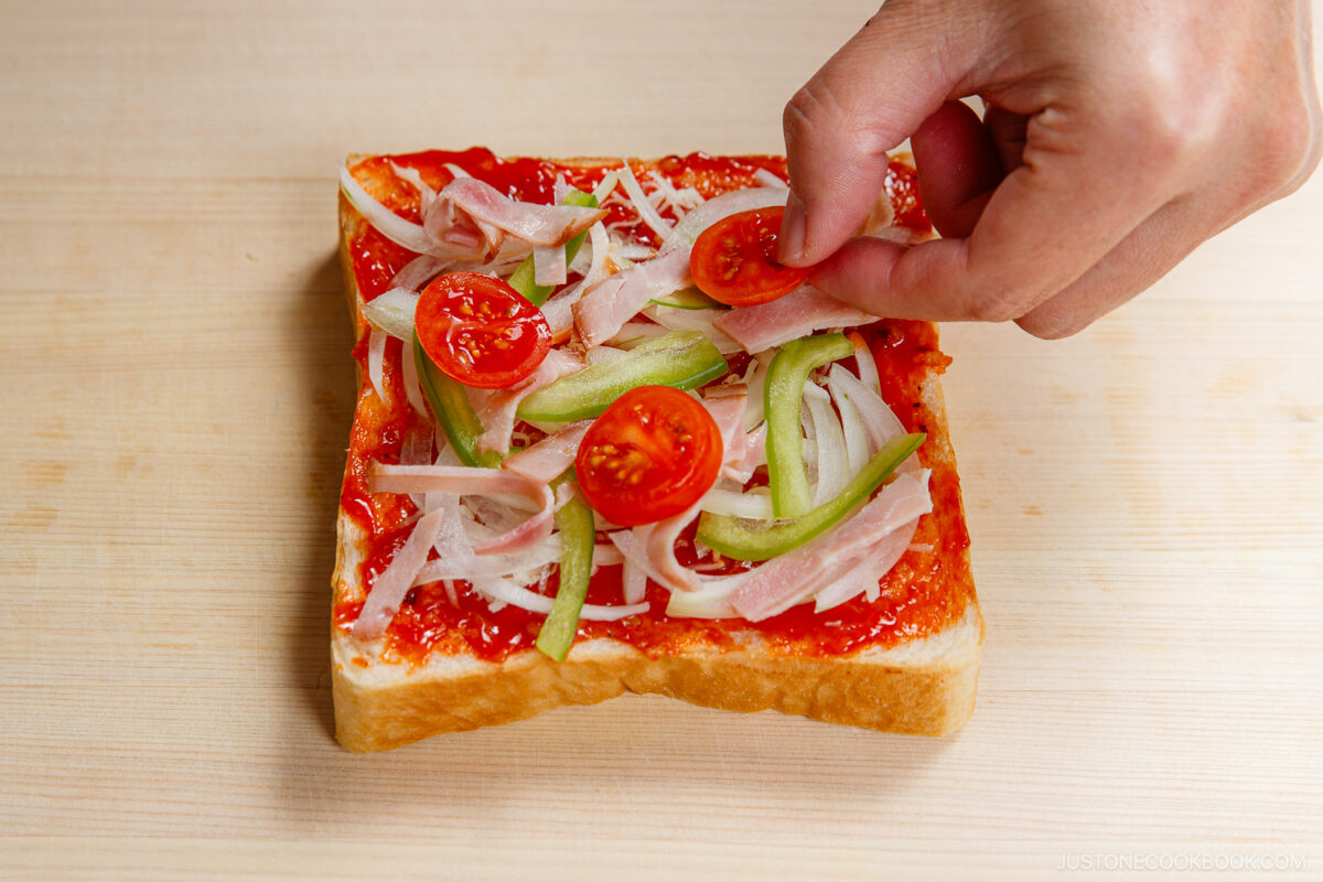 A hand places a slice of cherry tomato on a slice of bread topped with tomato sauce, green bell peppers, onions, ham strips, and other tomato slices, on a light wooden surface.
