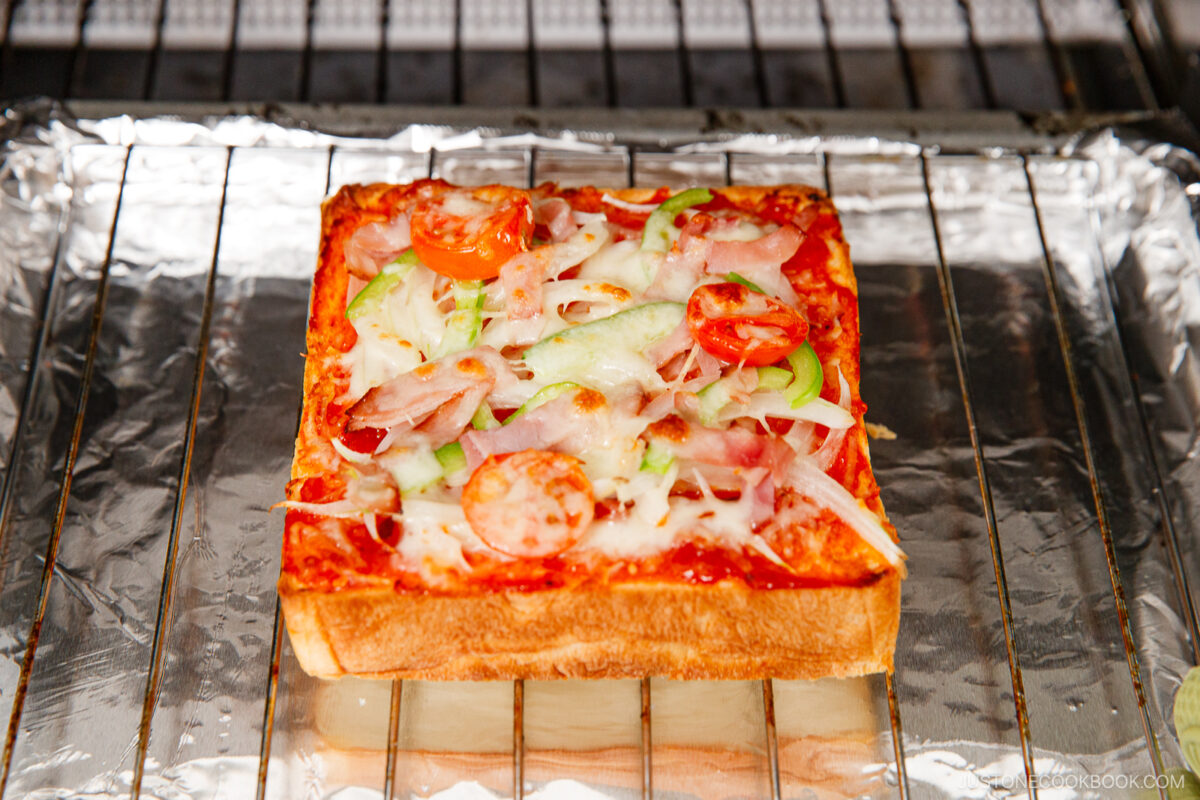 A thick slice of bread topped with melted cheese, tomato slices, green bell pepper, and onions, placed on a wire rack over foil in an oven.