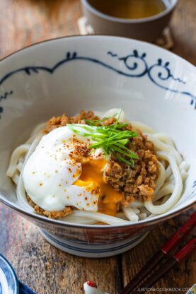 A bowl of udon noodles topped with seasoned ground meat, a poached egg with a runny yolk, sliced green onions, and a sprinkle of spices, served in a decorative bowl on a wooden table.