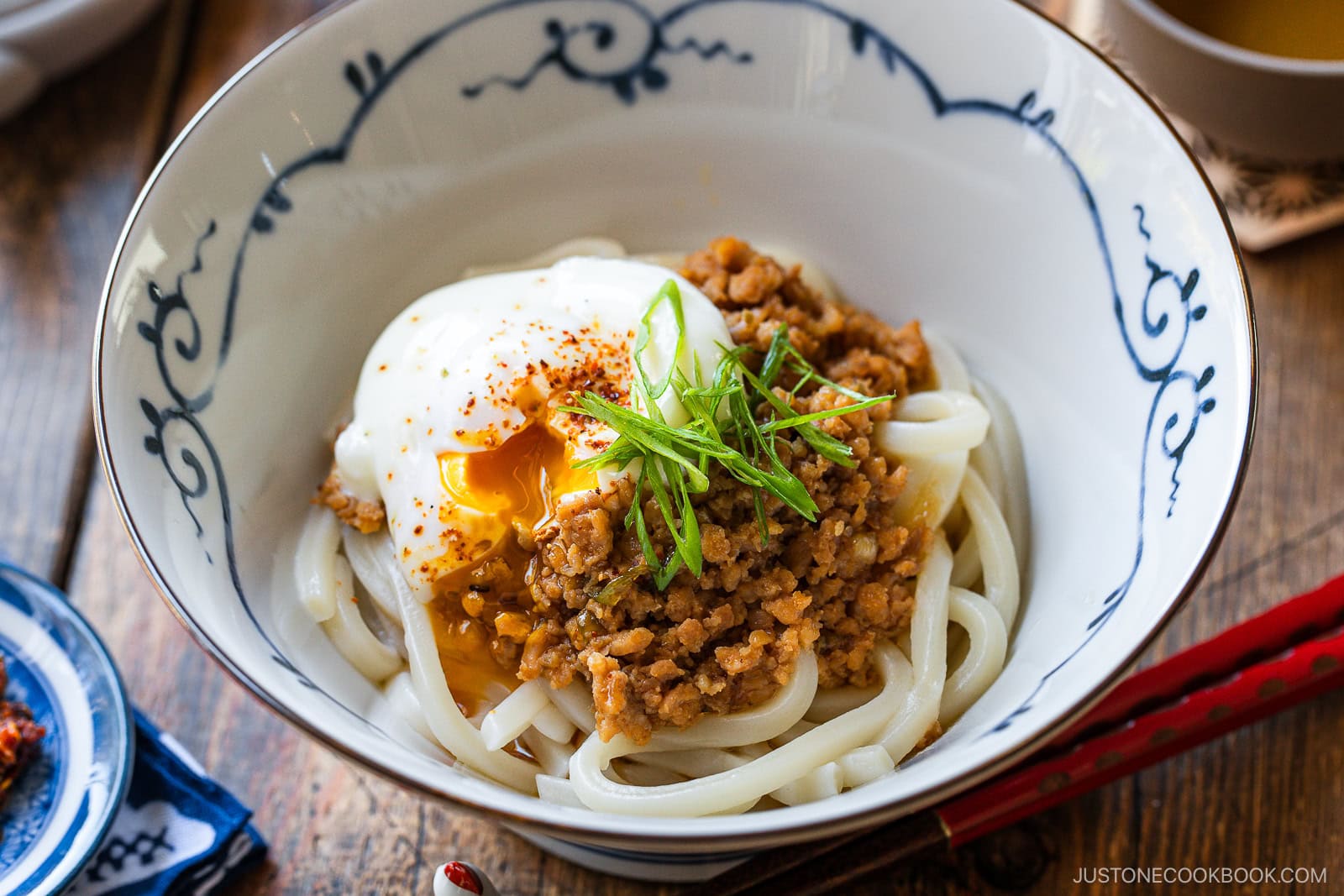 A bowl of udon noodles topped with minced meat, a soft-boiled egg with a runny yolk, sliced green onions, and a sprinkle of spices, served in a decorative white and blue bowl.