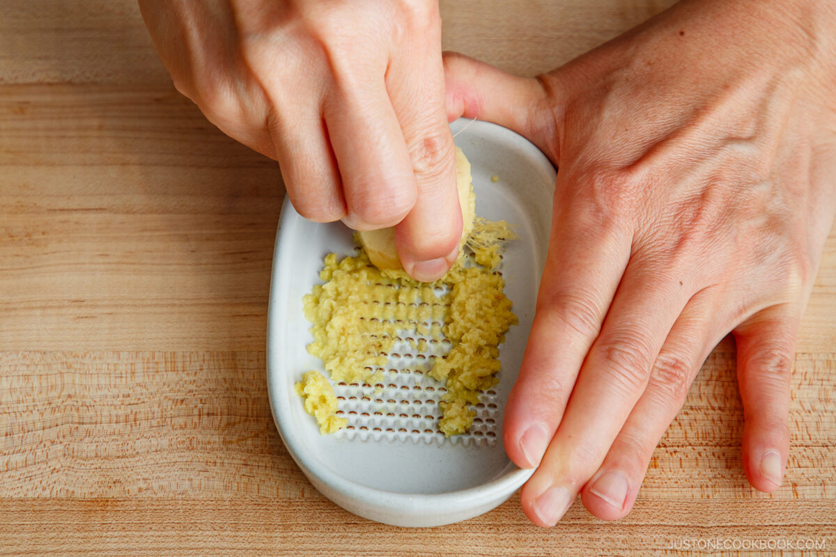 A person grates fresh ginger on a small white ceramic grater, holding the ginger with one hand and steadying the plate with the other, on a wooden surface.