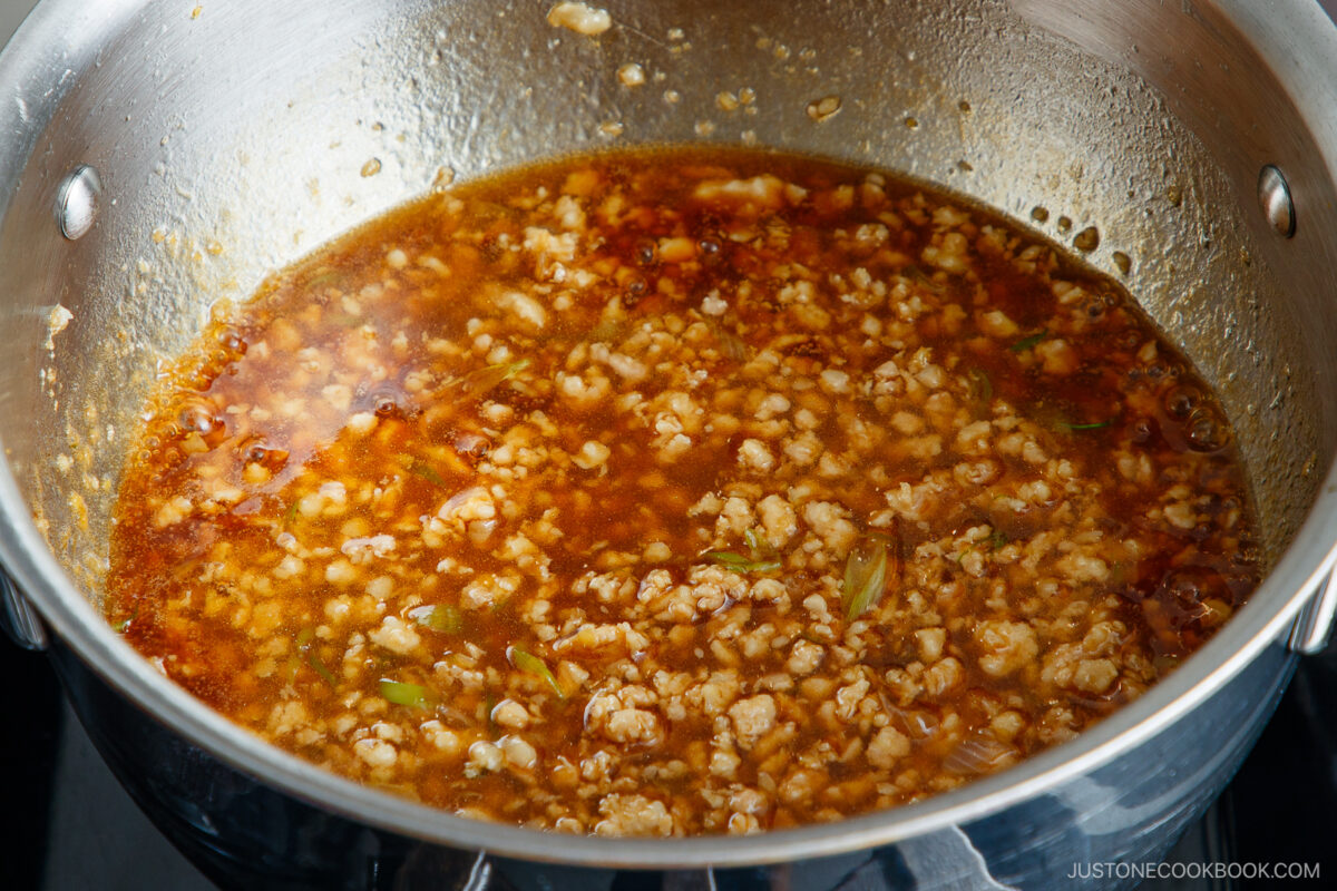 Ground meat simmering in a stainless steel pot with a rich, reddish-brown sauce, small bits of green herbs, and visible oil on the surface.