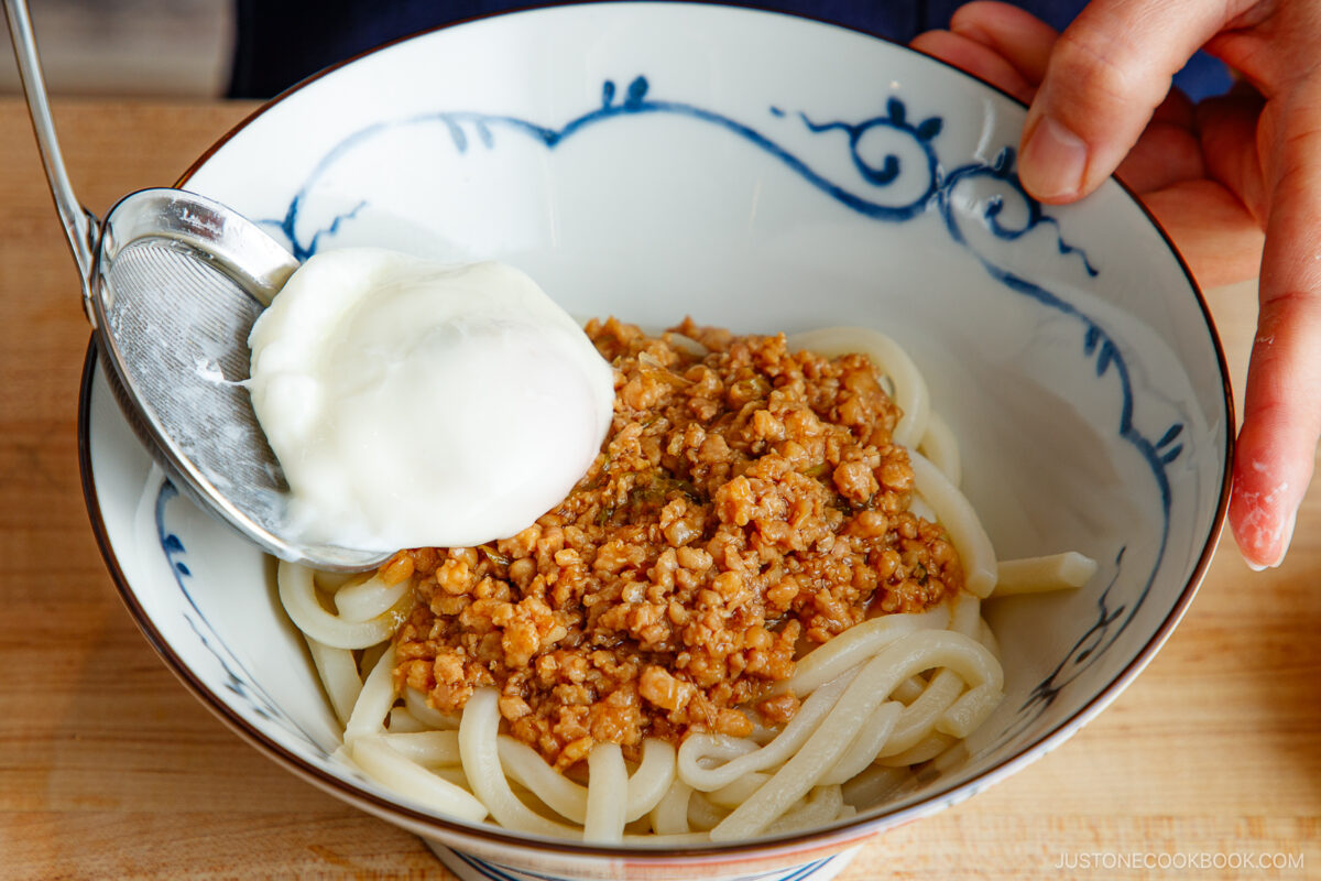 A bowl of udon noodles topped with minced meat sauce, with a hand holding a slotted spoon placing a poached egg on top. The bowl has a blue and white pattern.