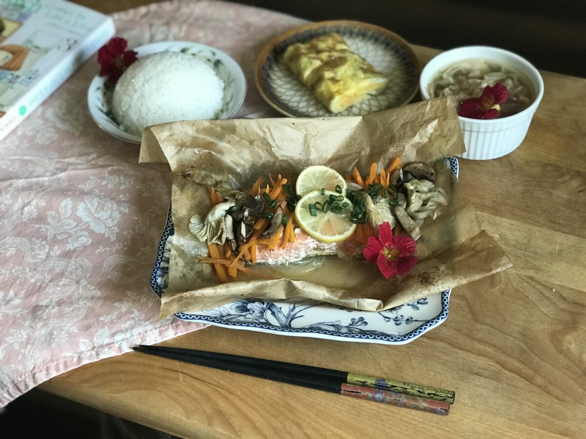 A wooden table set with a meal: baked fish with lemon, carrots, mushrooms, and herbs on parchment paper, a bowl of white rice, a rolled omelet, soup, and decorated with red flowers and chopsticks.