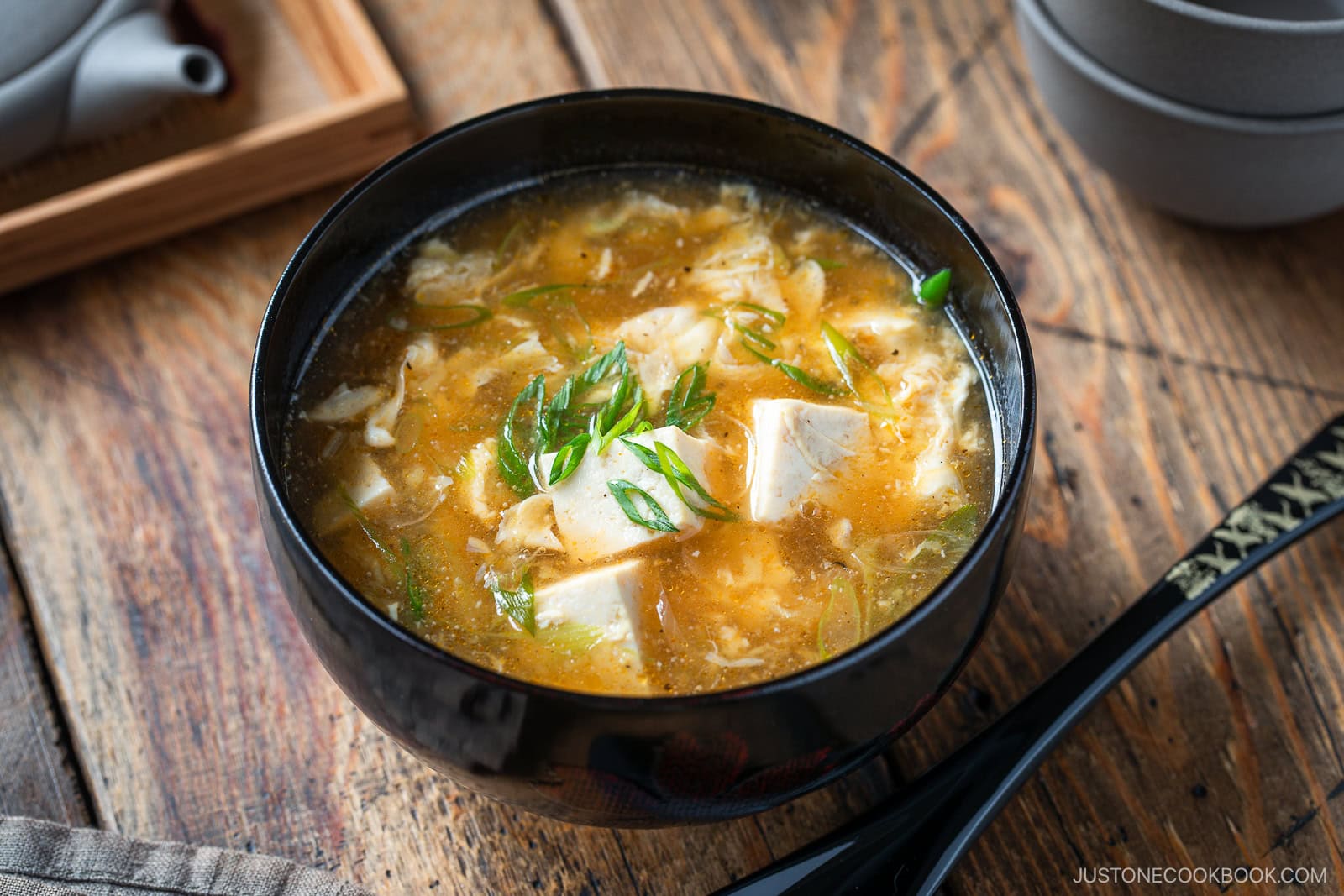 A bowl of hot and sour soup with tofu cubes, garnished with sliced green onions, sits on a wooden table next to a black spoon and teapot.