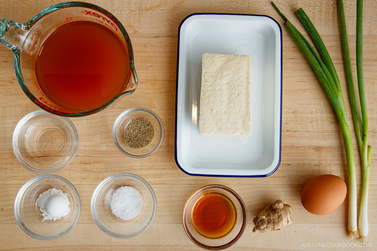 Overhead view of ingredients on a wooden surface: a glass measuring cup with broth, a block of tofu on a dish, green onions, an egg, ginger, soy sauce, pepper, cornstarch, and salt in small bowls.