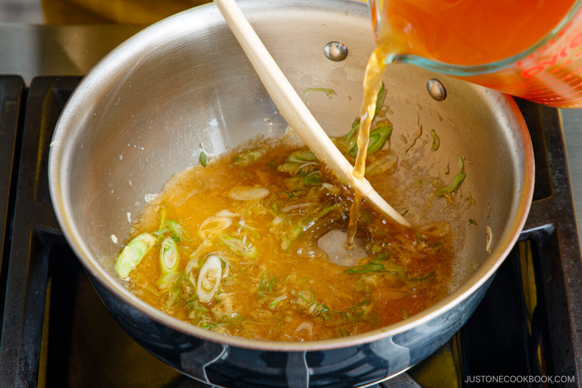 A hand pours broth from a measuring cup into a saucepan with sliced green onions and sauce, being stirred with a wooden spoon on a stovetop.