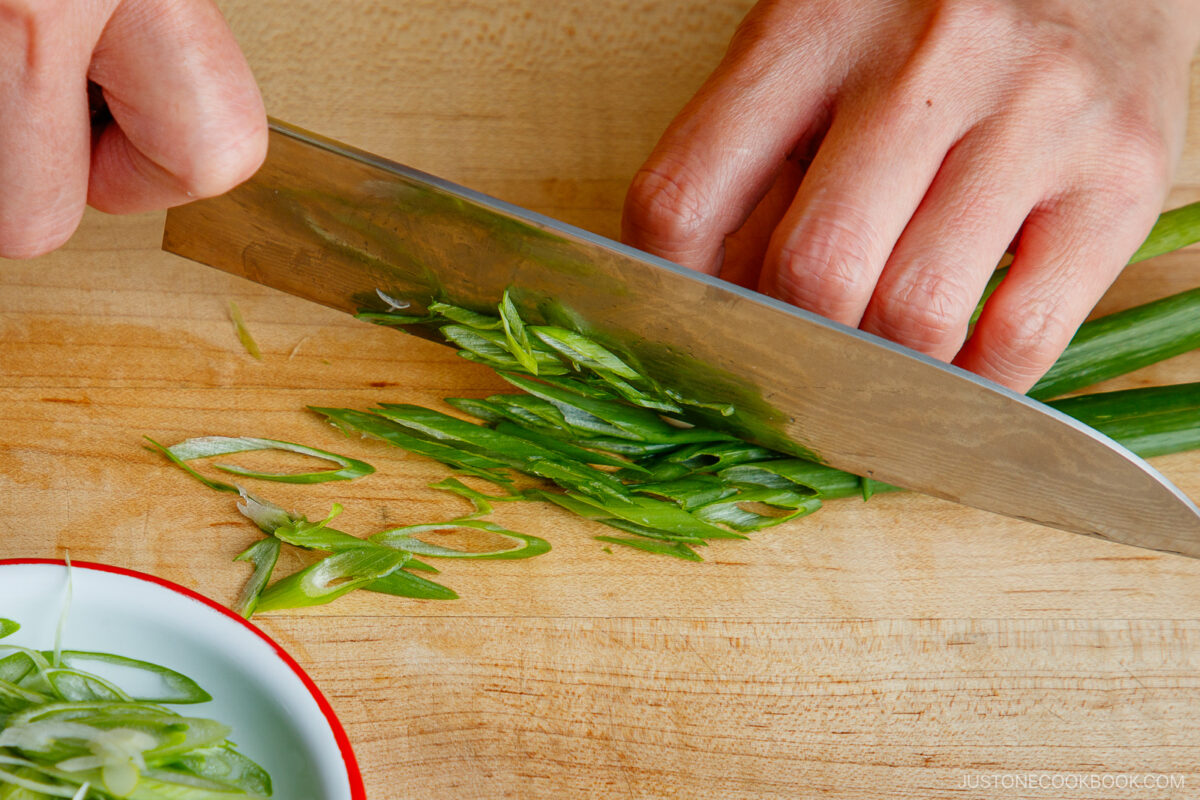 A person slices green onions on a wooden cutting board with a large knife; thinly cut green onion pieces are visible on the board and in a small dish.