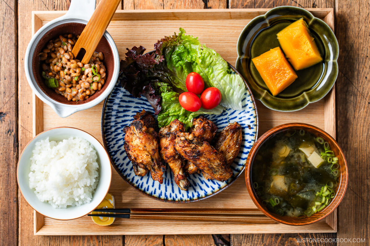 A Japanese meal set on a wooden tray with grilled chicken wings, lettuce, cherry tomatoes, steamed white rice, natto, miso soup with tofu and green onions, and simmered kabocha squash.
