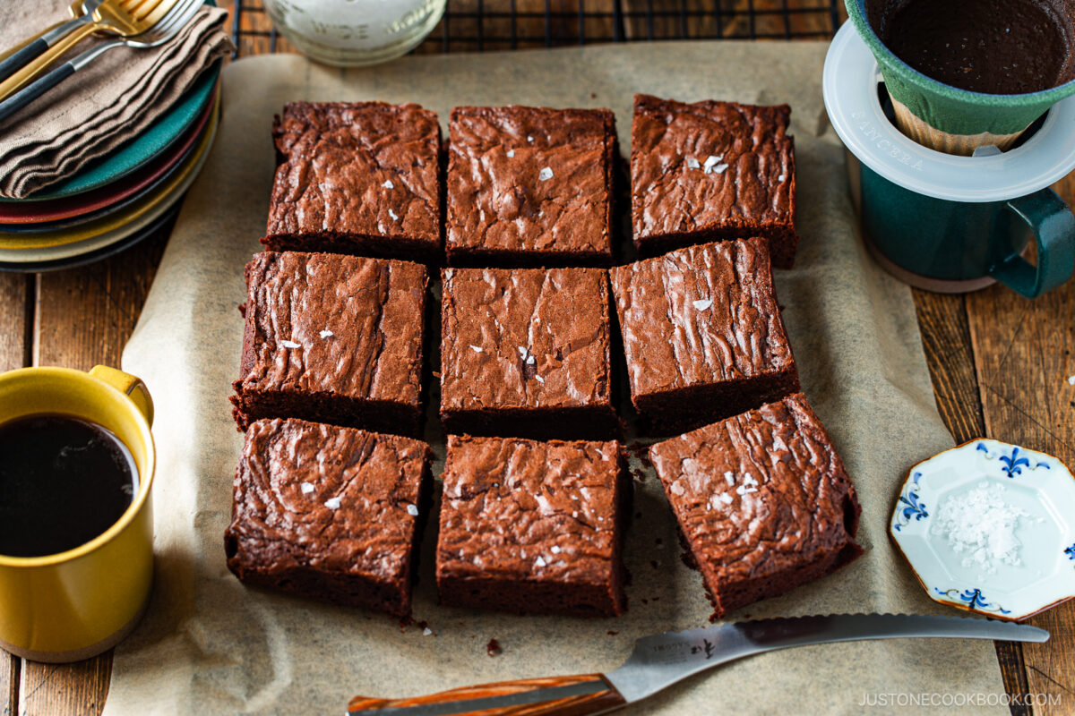 Nine square brownies sprinkled with sea salt are arranged on parchment paper, surrounded by a cup of black coffee, a knife, a stack of plates, and a small dish of salt on a wooden table.