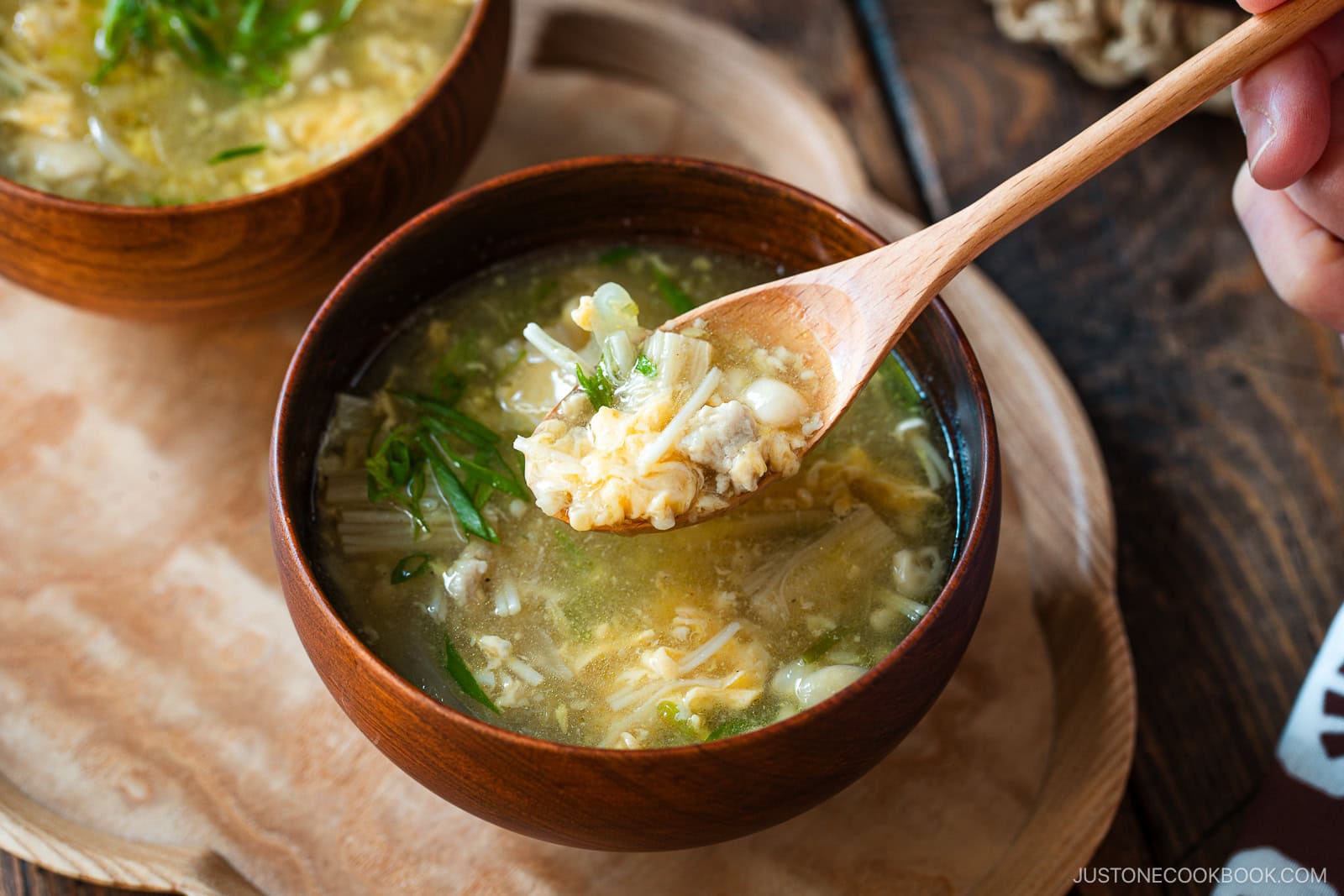 A close-up of a wooden bowl filled with egg drop soup containing vegetables and tofu; a hand holds a wooden spoon scooping up a bite from the bowl.