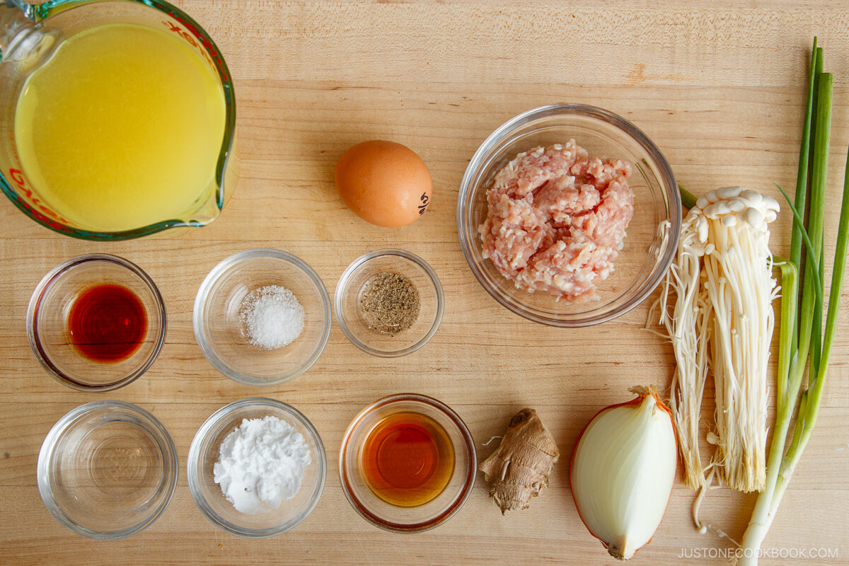 A top-down view of various ingredients on a wooden surface, including chicken broth, an egg, ground meat, enoki mushrooms, green onions, onion, ginger, and several small bowls of seasonings and sauces.