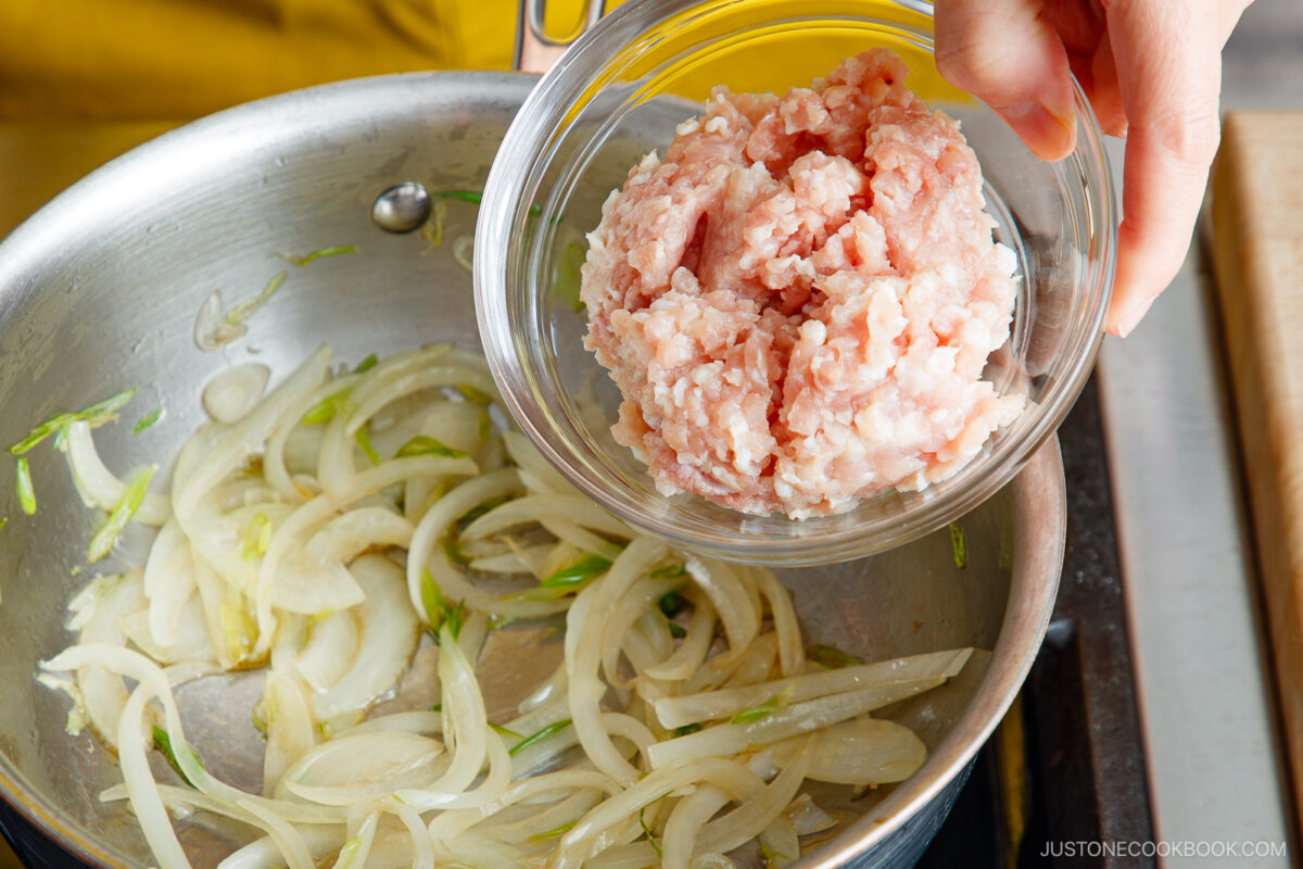 A hand holds a glass bowl of raw ground meat above a pan of sliced onions being sautéed on a stovetop, ready to add the meat to the pan.