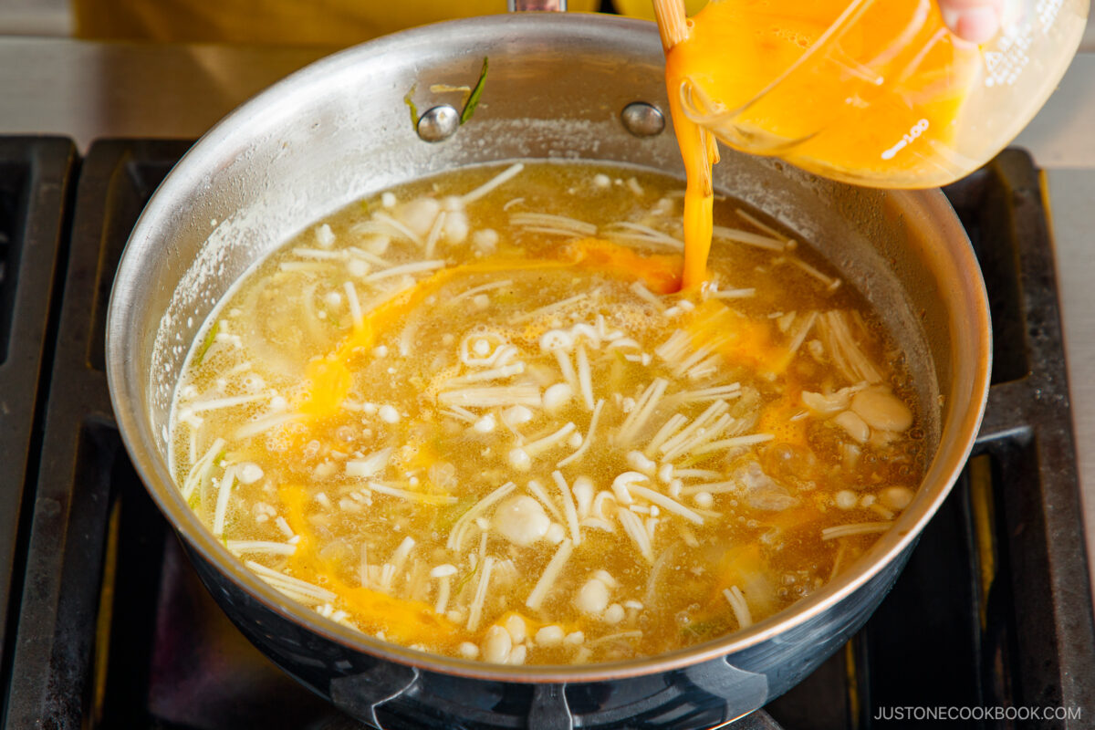 A hand pours beaten eggs from a measuring cup into a pot of soup with mushrooms and broth on a stovetop.