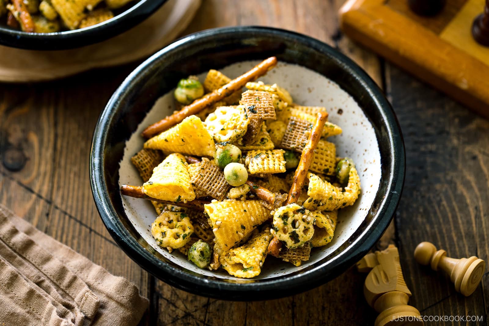 A bowl of snack mix featuring Chex cereal, pretzel sticks, rice crackers, and green wasabi peas sits on a wooden table next to a napkin and a chessboard, with warm natural lighting.