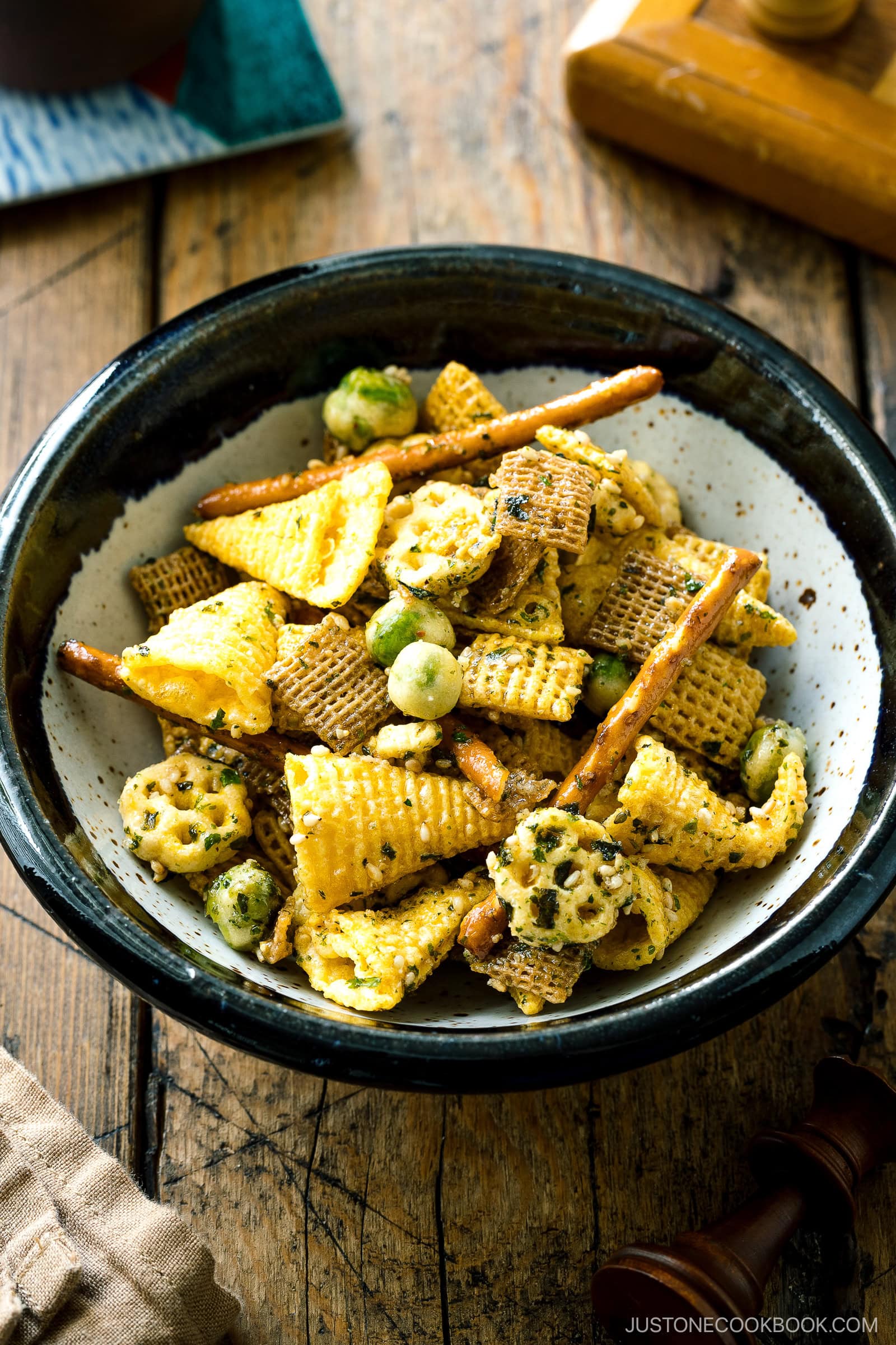 A bowl filled with a colorful snack mix, including rice crackers, pretzel sticks, wasabi peas, and various crunchy cereal pieces, sits on a wooden table.
