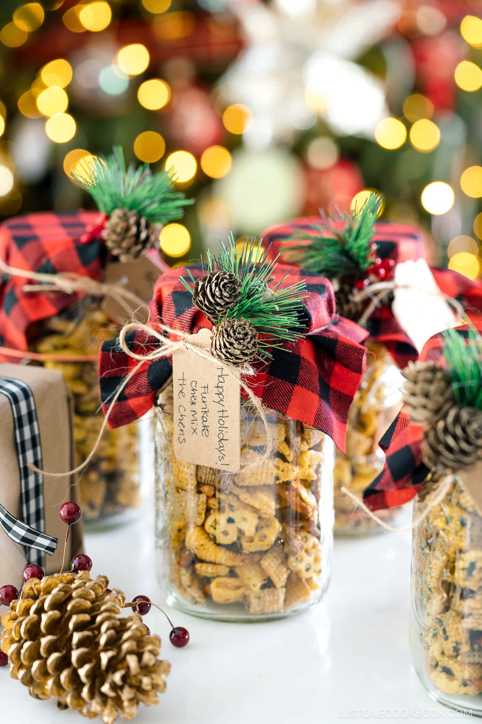 Glass jars filled with snacks are topped with red and black plaid fabric, pinecones, and greenery. Each jar has a holiday gift tag, with festive lights and decorations blurred in the background.